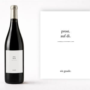May include: A black wine bottle with a silver cap and a white label. The label has the words "prost. auf di." and "ois guade." printed on it. Next to the bottle is a white card with the same text.