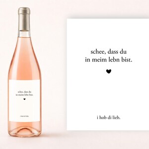 Puede incluir: Una botella de vino rosado con tapón plateado y etiqueta blanca. La etiqueta tiene el texto "schee, dass du in meim lebn bist." y "i hob di lieb." Una tarjeta blanca con el mismo texto y un símbolo de corazón está al lado.