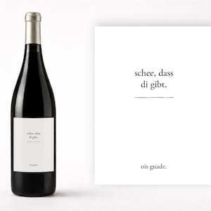 Puede incluir: Una botella de vino negra con tapón plateado y etiqueta blanca. La etiqueta tiene texto en idioma extranjero. Junto a la botella, una tarjeta blanca con texto en idioma extranjero.