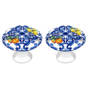 Peut inclure: Deux boutons de placard ronds avec une base blanche ornée d'un motif de volutes bleues et d'illustrations d'agrumes orange avec des feuilles vertes. Chaque bouton présente une surface supérieure lisse et arrondie et une base en métal argenté.