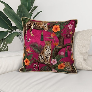 Pode incluir: Uma almofada quadrada decorativa com um fundo rosa vibrante apresentando uma cena da selva. O design inclui leopardos, flores de hibisco laranja e folhagem verde. A almofada tem uma borda dourada.