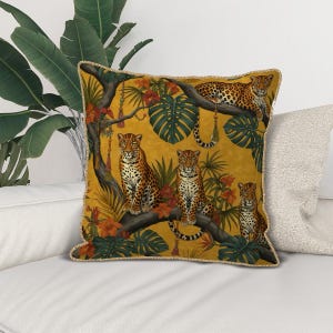 Könnte beinhalten: Quadratisches Zierkissen mit senfgelbem Hintergrund, das eine Dschungelszene mit Leoparden, tropischen Pflanzen und orangefarbenen Blumen zeigt. Das Kissen hat eine goldene Bordüre und liegt auf einem weißen Sofa.