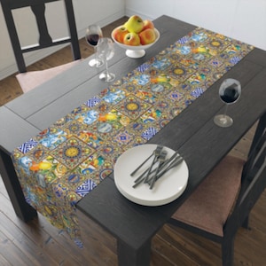 May include: A colorful table runner with a repeating tile pattern in shades of blue, yellow, and orange. The runner is placed on a dark wooden table, set with plates, silverware, and wine glasses. A bowl of apples sits in the center.