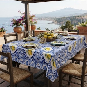 May include: A rectangular tablecloth featuring a blue and white tile-inspired pattern with yellow lemon motifs, seashells, and anchors. The table is set with white ceramic plates, clear wine glasses, and a woven basket filled with fresh lemons.