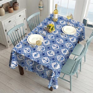 Peut inclure: Une table à manger rectangulaire recouverte d'une nappe blanche ornée d'un motif nautique bleu représentant des ancres, des poissons et des navires. La table est dressée avec deux assiettes blanches, deux verres à vin et un centre de table floral jaune.