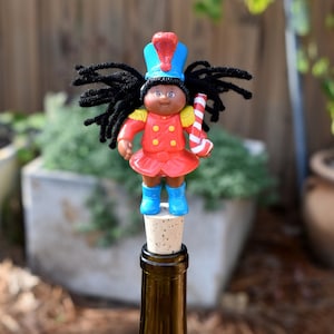 May include: A decorative bottle stopper featuring a doll-like figure in a red uniform, blue hat, and boots, holding a candy cane. The figure has black hair and is attached to a cork, sitting atop a green glass bottle.