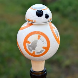 May include: A white and orange BB-8 droid bottle stopper. The spherical droid design features orange circular accents and black eye details. The bottle stopper is made of plastic and cork, and is inserted into a dark glass bottle.