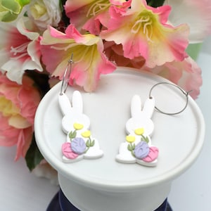 May include: White bunny-shaped hoop earrings with floral accents in yellow, pink, and purple. The earrings are displayed on a white pedestal, with a backdrop of pink and white flowers. The earrings are a charming accessory.
