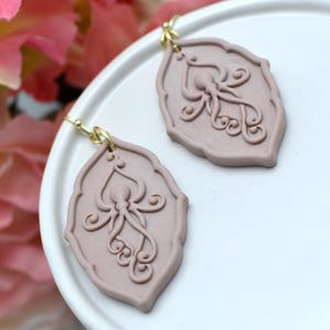 May include: A pair of mauve-colored dangle earrings featuring a raised octopus relief design on a scalloped, shield-shaped base. Each earring is attached to a gold-toned metal hook, showcasing a detailed, symmetrical cephalopod motif.