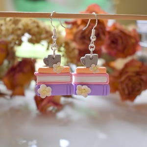 Puede incluir: Pendientes colgantes con pilas de libros coloridos, cada uno coronado con una taza gris y una pequeña flor blanca. Los libros son morados, rosas y naranjas, con ganchos plateados.