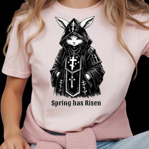 Puede incluir: Camiseta rosa claro con una ilustración en blanco y negro de un conejo con una túnica con capucha adornada con cruces. El texto "Spring has Risen" está impreso debajo. La camiseta está hecha de un material suave.