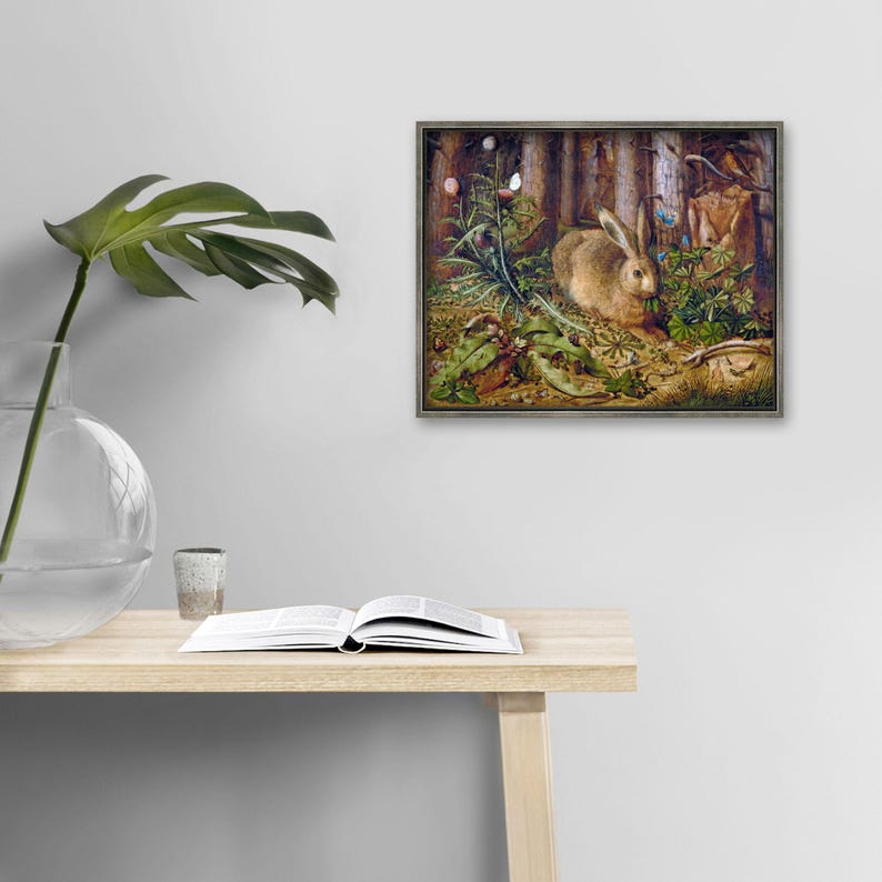 a painting of a rabbit in a forest setting, hanging on a wall above a wooden table with an open book and a glass vase.