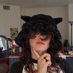 May include: A black, textured crochet hat with pointed ears and a wide brim. The hat is made of a soft, plush material. The hat is a unique accessory.