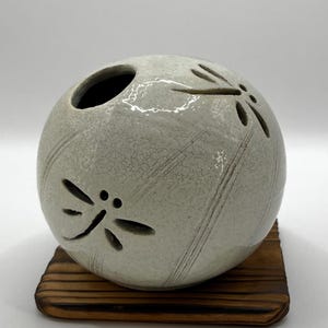 May include: A round, off-white ceramic decorative piece with dragonfly cutouts. The ceramic piece sits on a small, square, wooden base. The dragonfly cutouts are on the side of the ceramic piece.