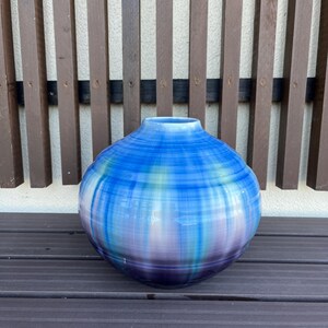 以下が含まれることがあります： ストライプ模様の丸い青と紫のセラミック花瓶。花瓶は幅広で球根状のボディと細い首を持っています。色が混ざり合い、グラデーション効果を生み出しています。花瓶は木製の表面に置かれています。