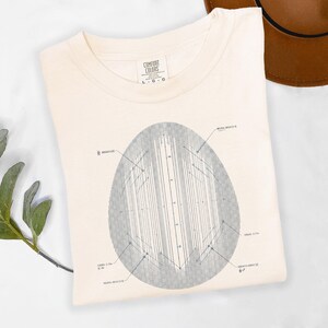 May include: A cream-colored t-shirt featuring a central graphic of a gray oval containing a technical diagram with vertical lines and various labels. The shirt has a crew neckline and a Comfort Colors brand tag at the inner collar indicating size large.