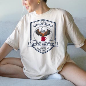 May include: A cream-colored t-shirt featuring a graphic of a red coffee mug with steam rising toward a window with a crescent moon. The text reads: EST. 1928 HERITAGE ROASTS TRADITIONAL COFFEE ROASTERY PREMIUM QUALITY HAND CRAFTED in blue ink.
