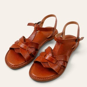 Op de afbeelding: Een paar bruine leren sandalen met een gevlochten bandontwerp over de voorvoet, een T-band in het midden en een verstelbare enkelband met een kleine metalen gesp. De sandalen hebben een platte zool en een gladde, gepolijste afwerking.