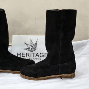 Puede incluir: Un par de botas de ante negro con suela plana. Las botas son altas, llegando a la mitad de la pantorrilla, y tienen un diseño sencillo y clásico. Las botas se exhiben sobre una superficie blanca con un logotipo en el fondo.