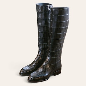 Peut inclure: Une paire de bottes hautes jusqu'au genou, de couleur gris anthracite foncé, avec une finition brillante et un motif texturé en peau de crocodile. Les bottes présentent un bout arrondi, un petit talon bloc et une fermeture éclair argentée.