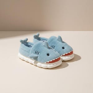 Puede incluir: Un par de zapatillas con forma de tiburón azul claro. Las zapatillas tienen suelas blancas y dientes rojos y blancos. Se ven las aletas y los ojos del tiburón. Estas zapatillas de felpa están diseñadas para la comodidad y la diversión.