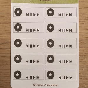 以下が含まれることがあります： 薄緑色の縁取りと筆記体で「Playlist」と書かれたステッカーシート。各ステッカーには黒い円とメディアコントロールアイコンが付いています。「Un carnet et une plume」というテキストが下にあります。