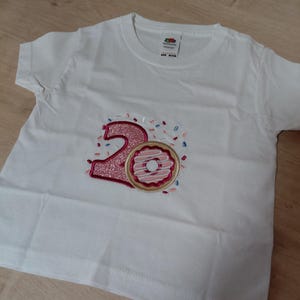 Op de afbeelding: Wit T-shirt met een roze glitter nummer "2" en een donut ontwerp. De donut heeft roze glazuur en sprinkles. Het shirt is gemaakt van een zacht materiaal en is geschikt voor kinderen.