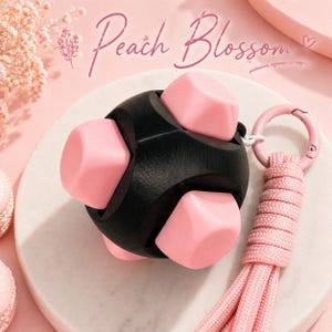 May include: A black and pink fidget toy with the text "Peach Blossom". The toy has a spherical shape with pink cube-shaped pieces. It includes a pink keychain with a braided cord.