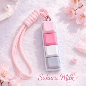 Puede incluir: Un llavero rectangular con cuatro botones cuadrados en tonos rosa, blanco y gris. El llavero tiene un cordón rosa y el texto "Sakura Milk" en una fuente cursiva.