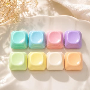 Könnte beinhalten: Acht pastellfarbene, quadratische Objekte mit einer abgerundeten Vertiefung in der Mitte. Die Farben umfassen Blau-, Rosa-, Lila-, Gelb- und Orangetöne. Die Objekte sind in zwei Reihen zu je vier Stück auf einer weißen, geriffelten Oberfläche angeordnet.