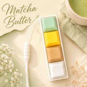 Könnte beinhalten: Ein rechteckiges weißes Makro-Tastenfeld mit vier quadratischen Tasten in Hellgrün, Gelb, Hellbraun und Weiß, verbunden durch ein weißes Kabel. Der Text "Matcha Butter" ist in einer grünen Schreibschrift auf dem hellen Hintergrund gedruckt.