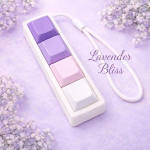 Könnte beinhalten: Ein rechteckiges, weißes Tastaturzubehör mit vier quadratischen Tasten in Lila, Rosa und Weiß. Ein weißes Handgelenkband ist befestigt. Der Text "Lavender Bliss" ist kursiv geschrieben.