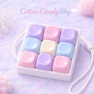 Könnte beinhalten: Eine weiße Schale mit neun quadratischen Objekten in Pastellfarben. Die Farben umfassen Rosa, Lila, Blau und Creme. Der Text "Cotton Candy Sky" steht in Schreibschrift über der Schale. Ein weißes Band ist befestigt.
