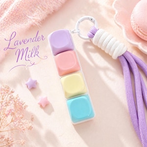 May include: A keyring featuring four square, pastel-coloured keys in lavender, pink, yellow, and blue, attached to a white ring with a purple cord. The text Lavender Milk is written in a cursive script on the left side of the keyring.