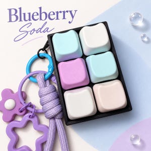 May include: A keychain featuring a black rectangular tray holding six square keycaps in pastel blue, white, and purple. Attached is a purple braided cord, a purple flower charm, a purple star-shaped ring, and a blue ring. Text reads Blueberry Soda.