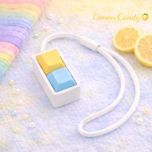 Könnte beinhalten: Ein weißer, rechteckiger Anhänger mit gelben und blauen Tastenkappen, befestigt an einem weißen Schlüsselband. Das Bild enthält den Text "Lemon Candy" und zwei Zitronenscheiben. Der Hintergrund ist ein sanftes Pastellfarbschema.