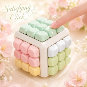 May include: A cube-shaped fidget toy with a white frame and rows of soft-coloured square buttons in pastel green, pink, yellow, and light blue. The text Satisfying Click is printed in a decorative script font above the cube.