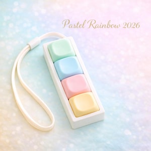 May include: A white rectangular accessory with four pastel-coloured square buttons: green, blue, pink, and yellow. The accessory has a white strap attached. The text "Pastel Rainbow 2026" is written in gold script.