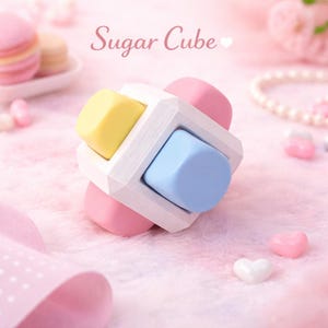 Puede incluir: Un juguete en forma de cubo con colores pastel, amarillo, azul y rosa. El juguete es blanco y tiene la inscripción "Sugar Cube" en cursiva. El fondo es rosa claro, con caramelos en forma de corazón y un collar de perlas.