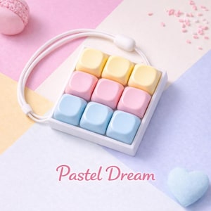 Könnte beinhalten: Ein quadratisches Makro-Pad mit neun Tasten, einem weißen Rahmen und pastellfarbenen Tastenkappen in Gelb, Rosa und Blau. Das Gerät verfügt über eine weiße Handschlaufe an der Oberseite und den Schriftzug "Pastel Dream" in rosa Schreibschrift.