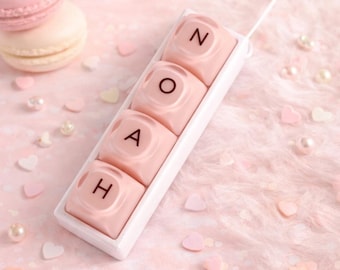 May include: A pink personalized keychain spelling out the name "NOAH" in individual letter blocks. The keychain is in a white rectangular case, with a white string attached. The background is a soft pink with scattered pearls, heart-shaped confetti, and a macaron.