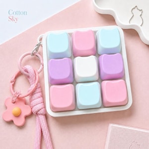 May include: A square white frame containing nine pastel-coloured keycaps in light blue, pink, white, and purple. Attached to the frame is a pink keychain with a braided cord and a flower-shaped charm with a yellow centre. The text Cotton Sky is visible.