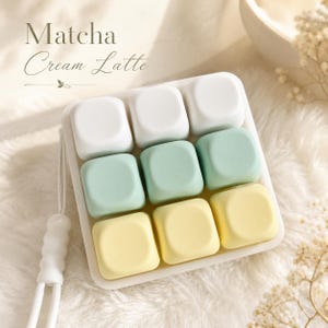 Puede incluir: Una bandeja cuadrada que contiene nueve piezas con forma de cubo redondeado dispuestas en tres filas de tres, en colores blanco, verde claro y amarillo pálido. El texto Matcha Cream Latte aparece impreso arriba a la izquierda con una hoja.