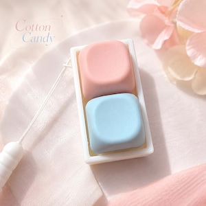 Puede incluir: Primer plano de un pequeño recipiente rectangular blanco que contiene dos objetos cuadrados. Uno es rosa claro, el otro azul claro. El texto "Cotton Candy" está en la esquina superior izquierda. El fondo es rosa suave.