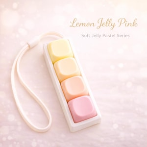 Può includere: Una custodia bianca rettangolare contiene quattro blocchi quadrati dai colori pastello: giallo, arancione chiaro, pesca e rosa. È attaccato un cordino bianco. Il testo recita "Lemon Jelly Pink" e "Soft Jelly Pastel Series."