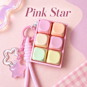 May include: A rectangular pink keyring featuring six pastel-coloured square keycaps in shades of pink, peach, yellow, and light green. The accessory includes a pink braided cord, a pink metal clasp, and a translucent pink star charm.