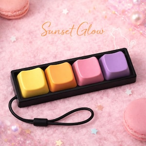 May include: A black tray holds four colorful keyboard keycaps in yellow, orange, pink, and purple. The keycaps are arranged in a row, with a black wrist strap attached. The text "Sunset Glow" is written in cursive above the keycaps.