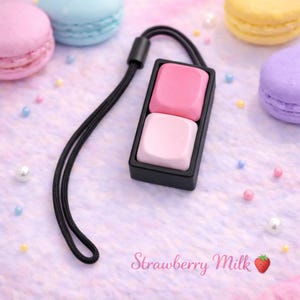 Könnte beinhalten: Ein schwarzes rechteckiges Accessoire mit zwei rosa Quadraten, befestigt an einer schwarzen Kordel. Der Text "Strawberry Milk" ist in rosa Schrift geschrieben. Pastell-Macarons und kleine dekorative Perlen befinden sich im Hintergrund.