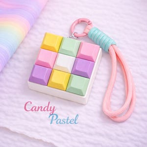以下が含まれることがあります： パステルカラーのキーホルダー。黄色、緑、ピンク、紫、白の正方形のキーキャップが9つ、3x3のグリッド状に配置されています。キーホルダーは白いフレーム、ピンクの留め金、ピンクとターコイズのストラップが付いています。「Candy Pastel」の文字が見えます。