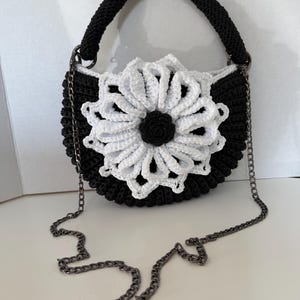 May include: A black and white crochet handbag with a flower design. The bag features a black handle and a silver chain strap. The flower is white with a black center, adding a striking contrast to the bag's design. The bag is approximately 8 inches wide.